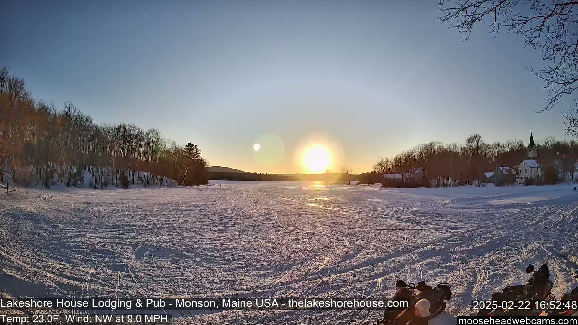 A frozen lake at sunset, under a clear sky, shows snowmobile tracks across the ice, with trees and buildings on the far shore.