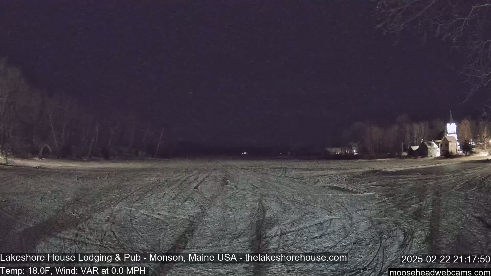 A snow-covered field at night, under a dark sky, with a few buildings and trees visible in the distance.