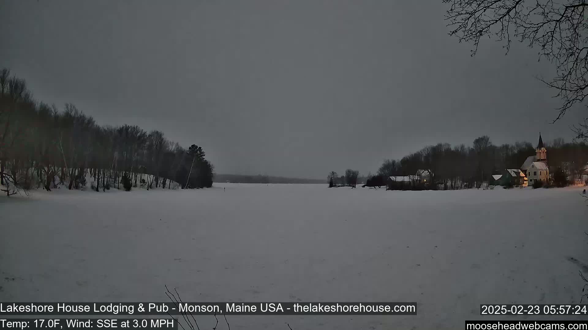 A snow-covered frozen lake is seen under an overcast sky, with a few buildings and trees visible on the far shore.