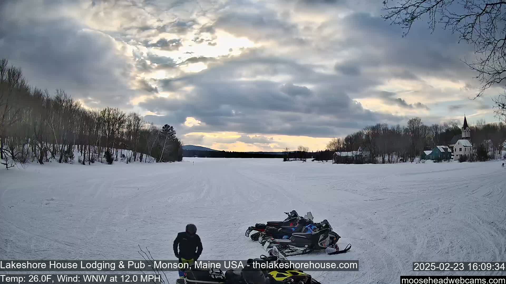 A person stands near several snowmobiles parked on a snow-covered frozen lake under a partly cloudy sky, with a village visible in the distance.