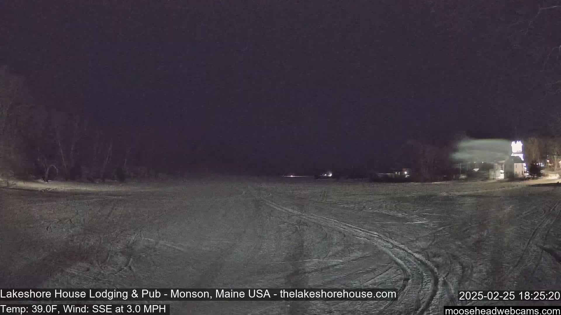A snow-covered, flat expanse stretches from the foreground to the background under a dark night sky, with a few dimly lit buildings visible in the distance and a slight wind.