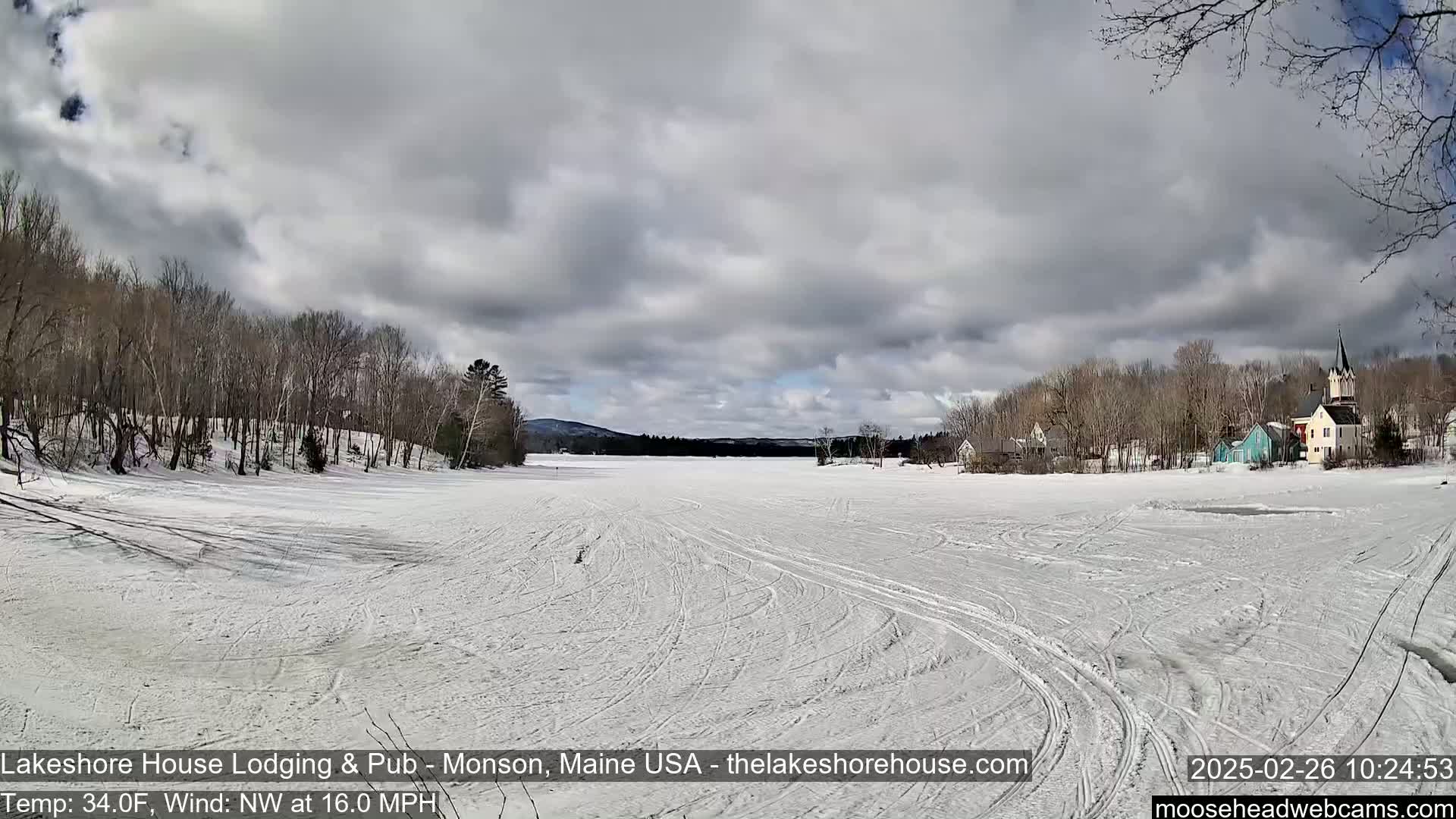 A frozen lake stretches to a town of houses and trees under a cloudy sky with a northwest wind.