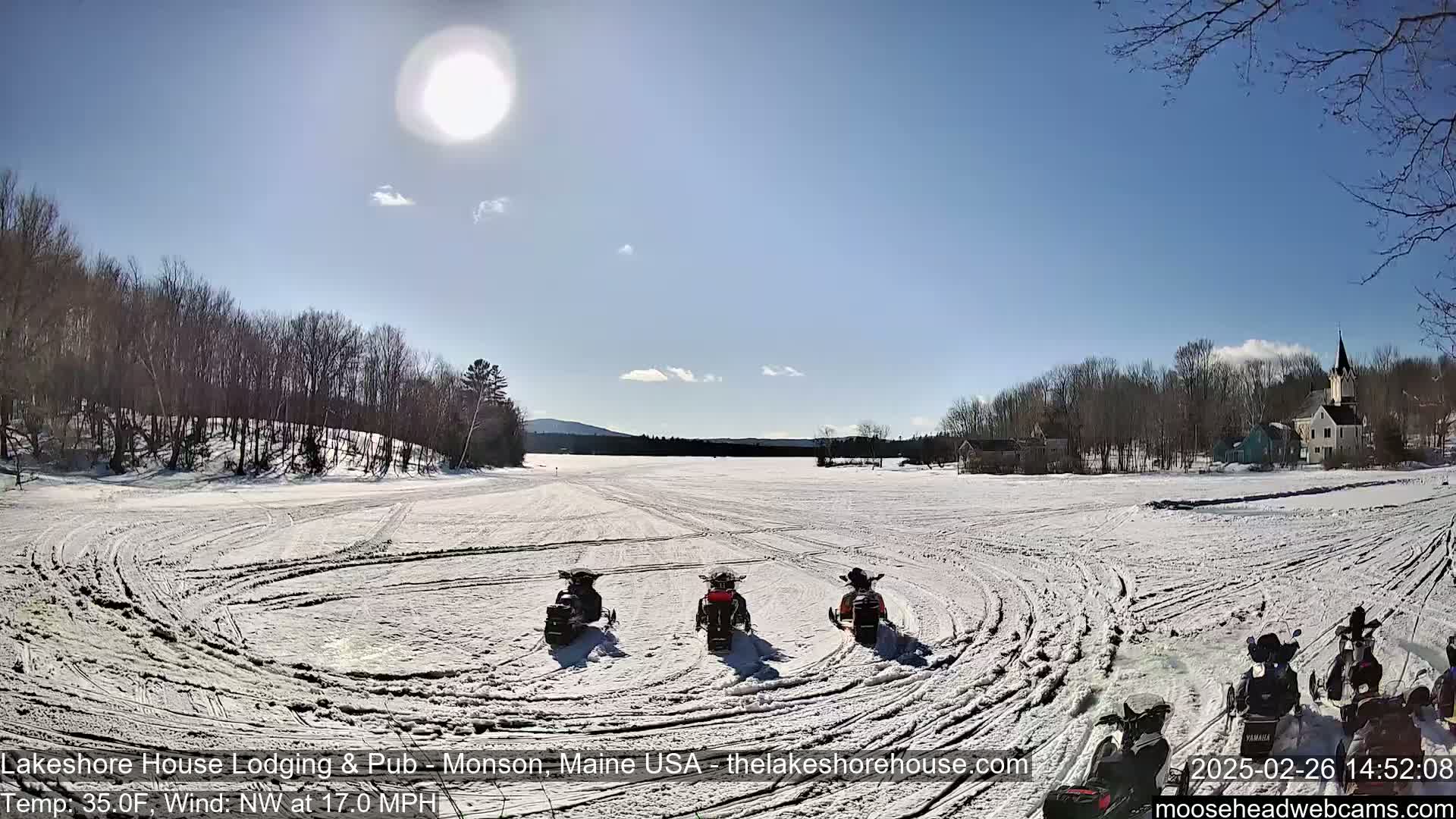 Three snowmobiles are parked on a snow-covered expanse of land under a bright, sunny sky with a light wind.