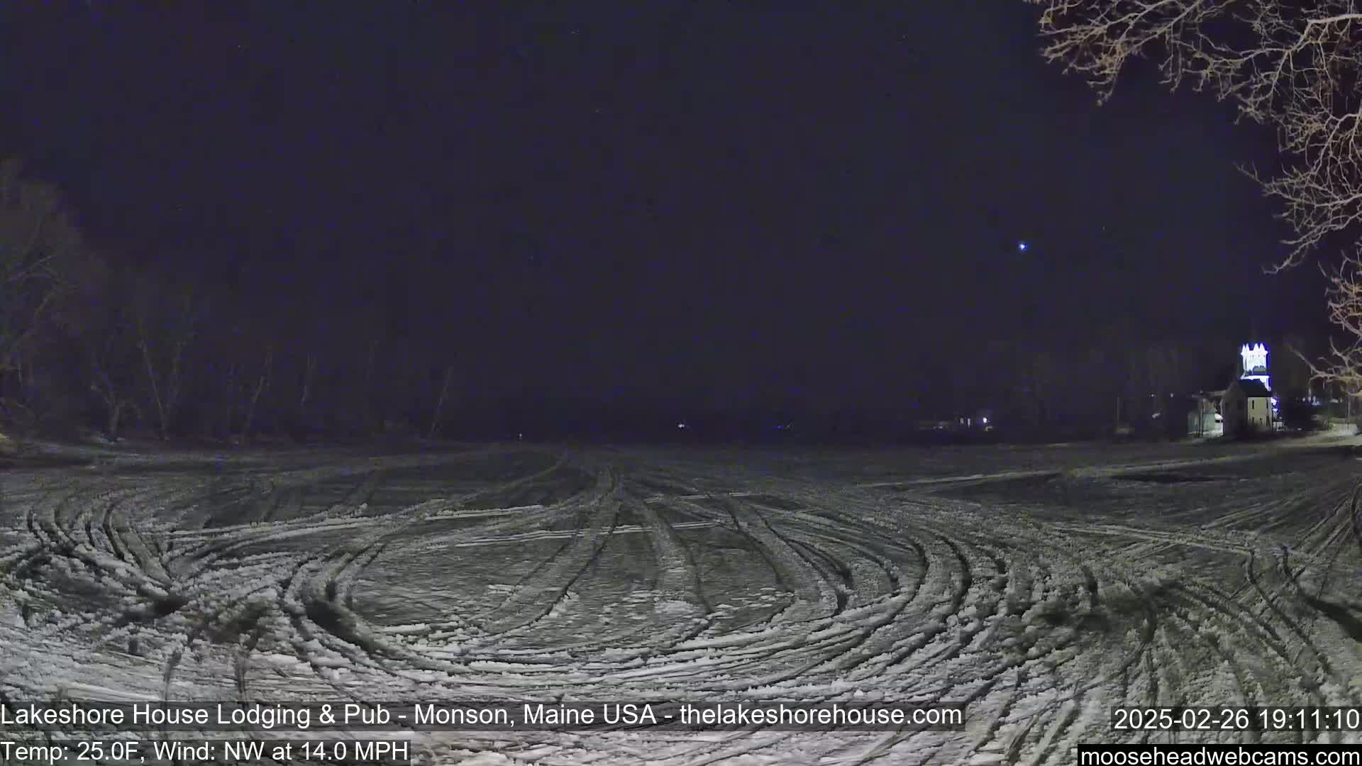 A snow-covered, dark field with tire tracks under a clear night sky, with a building visible in the distance and a northwest wind of 14 mph at 25 degrees Fahrenheit.