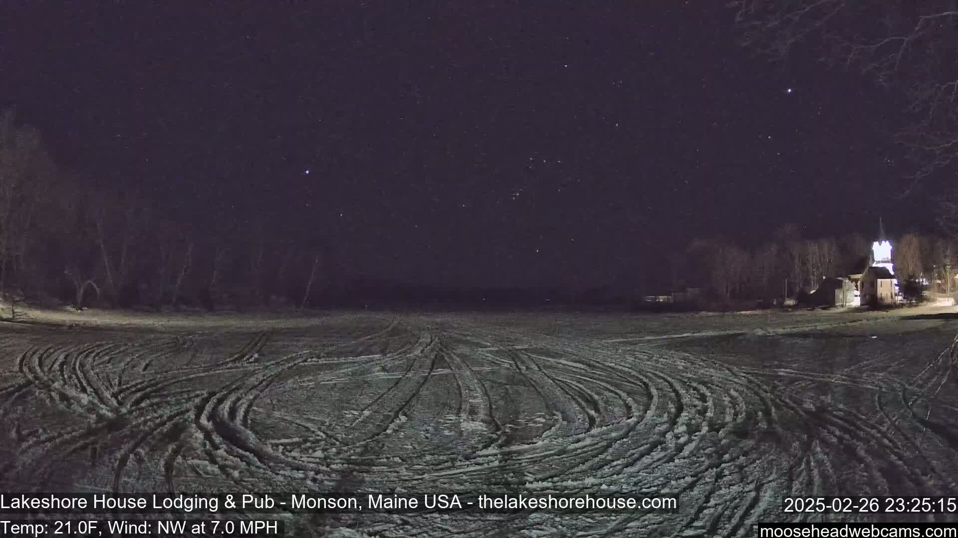 A snow-covered field with tire tracks is visible under a clear, starry night sky, with a building and trees in the distance.