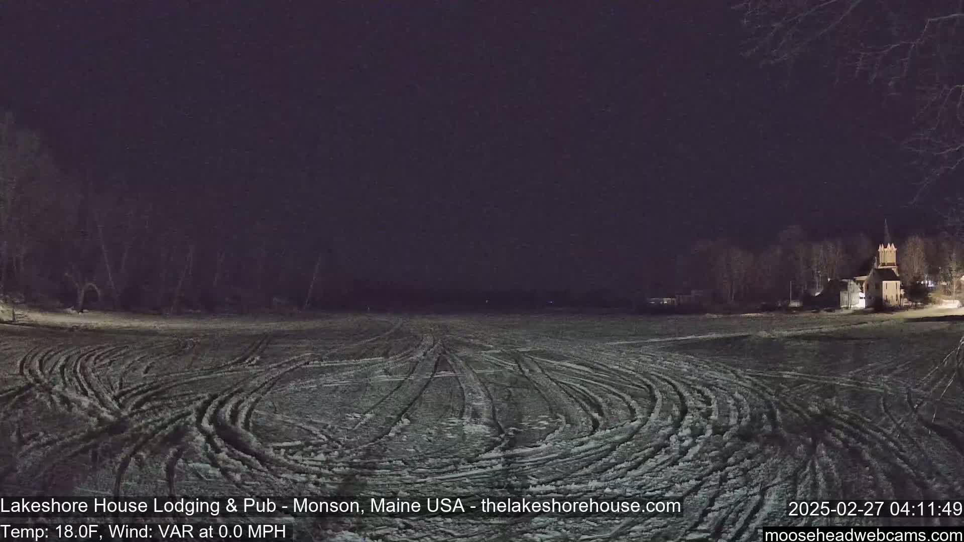 A snow-covered field with tire tracks is visible at night under a dark sky, with a building and trees in the distance.