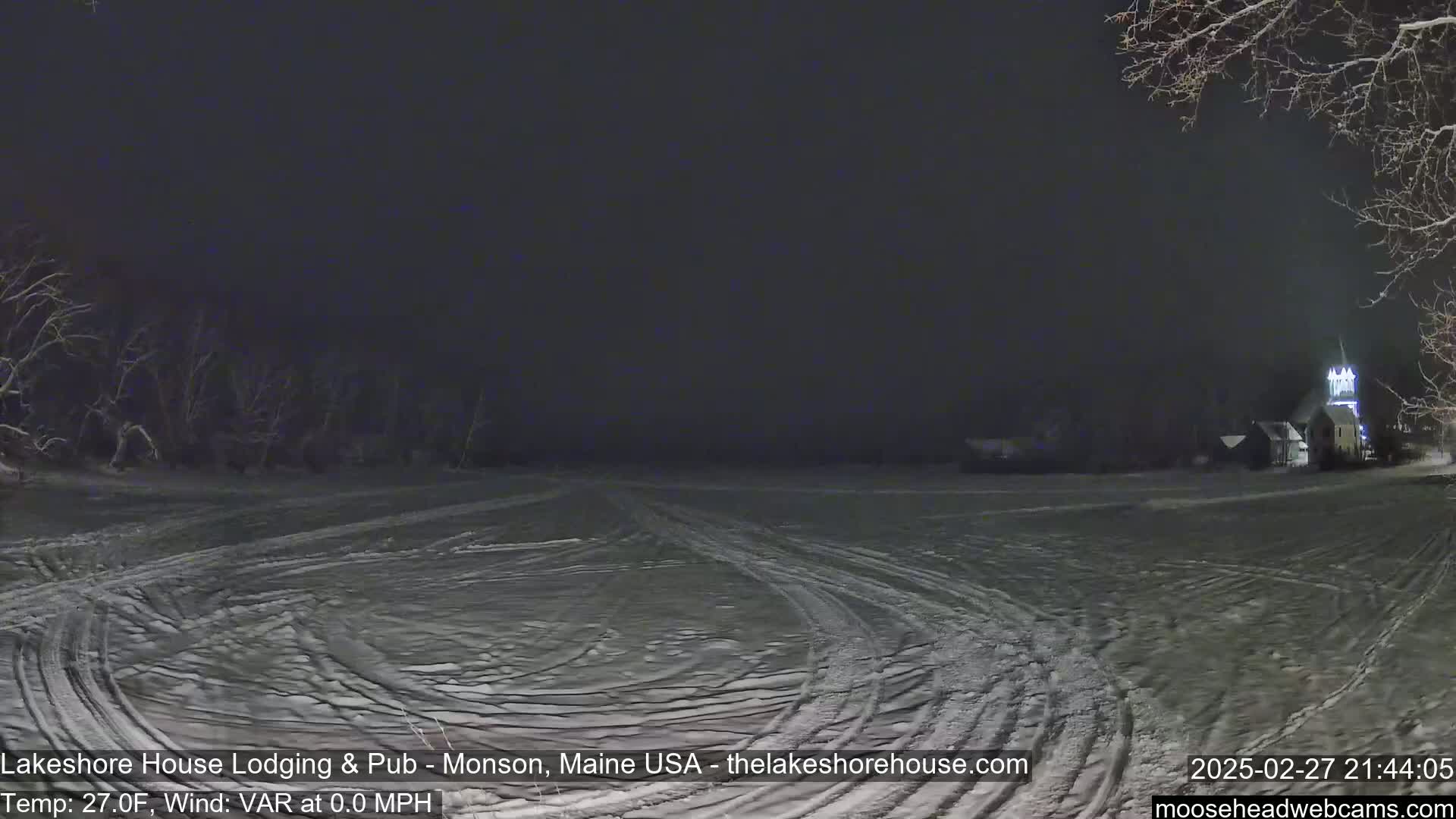 A snow-covered field at night with tire tracks and a building with lights visible in the distance under a dark sky.