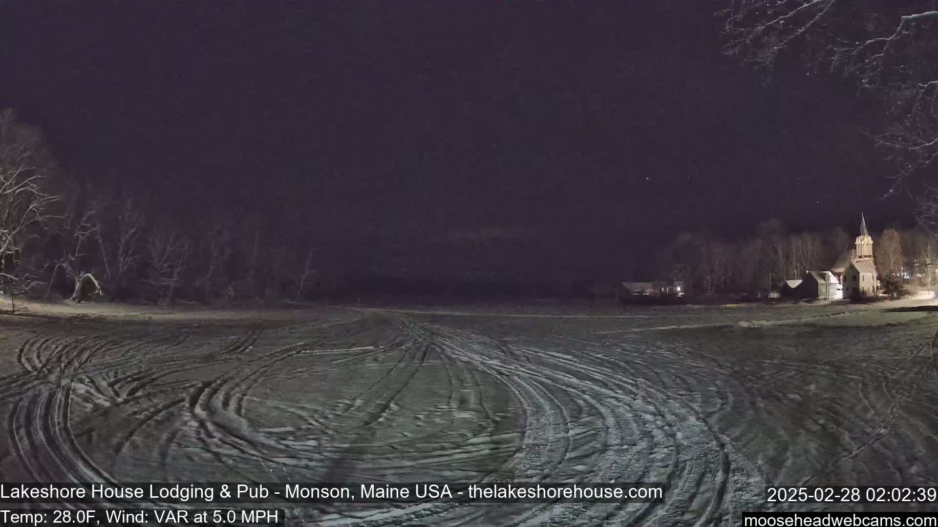 A snow-covered field at night, with tire tracks visible, and a church steeple in the distance under a dark sky.
