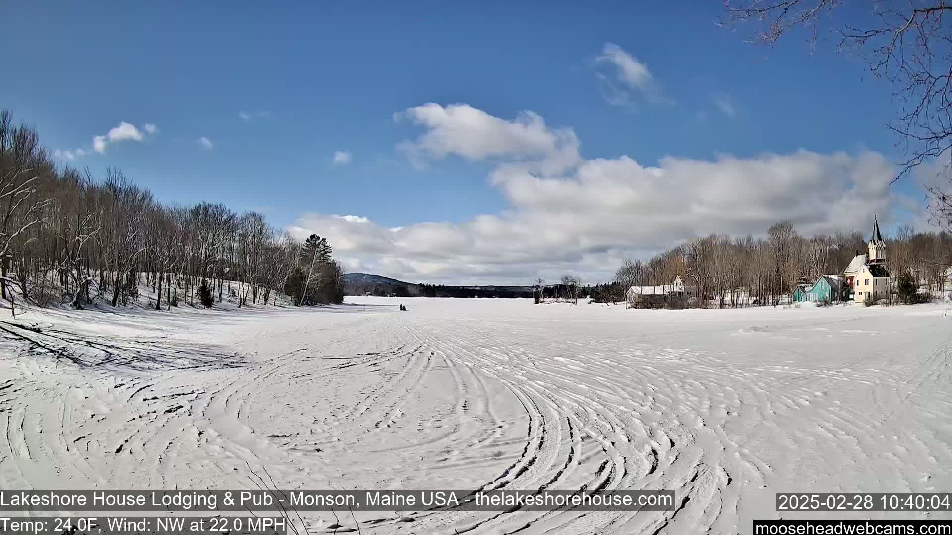A snow-covered frozen lake stretches out to a small village under a partly cloudy blue sky, with a strong northwest wind.