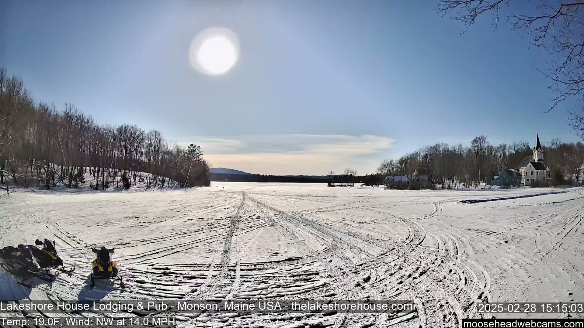 Two snowmobiles are parked on a snow-covered frozen lake under a bright, sunny sky, with a small town and trees in the background.