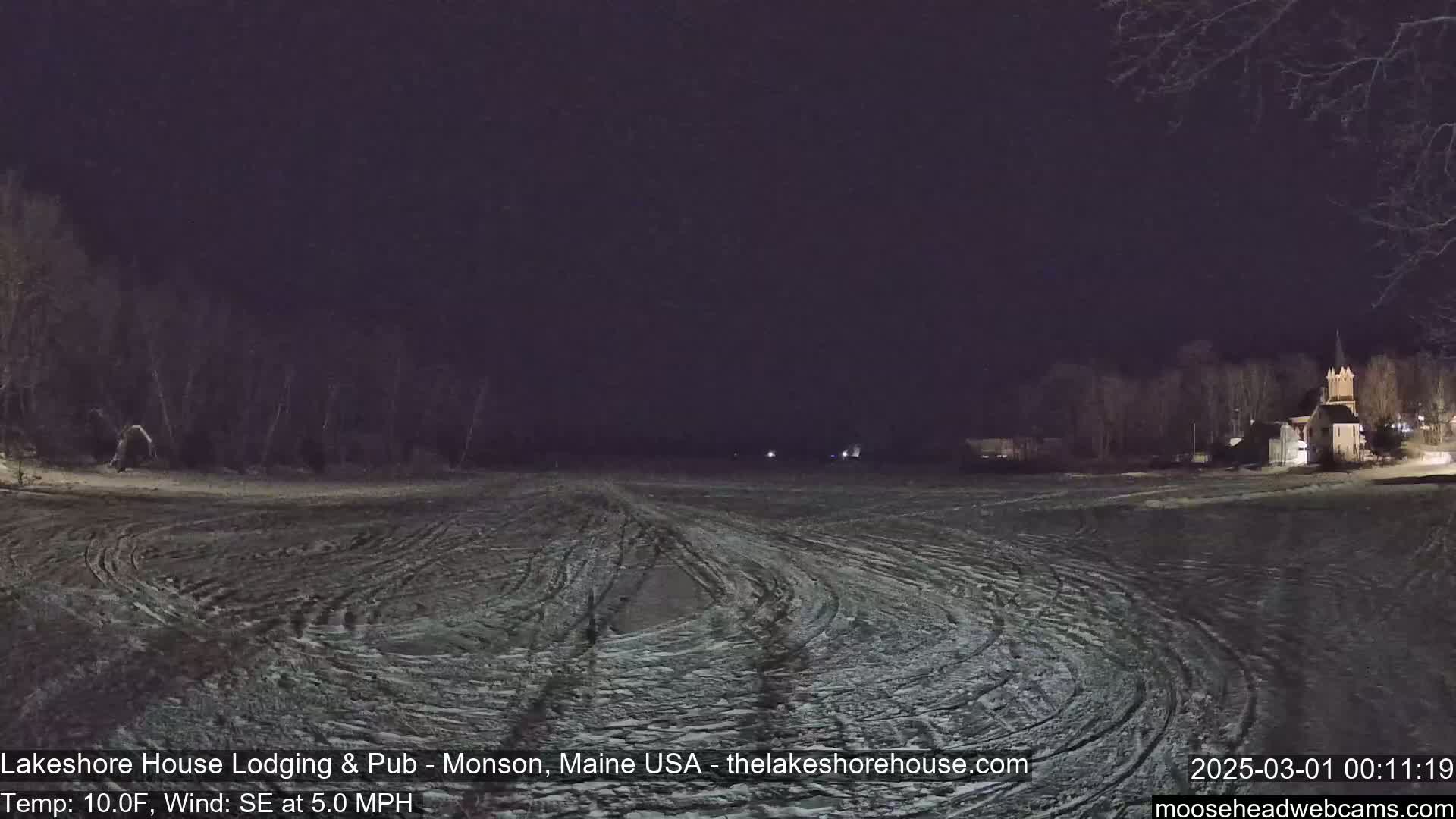 A snow-covered field at night, with a building and trees visible in the distance under a dark sky.