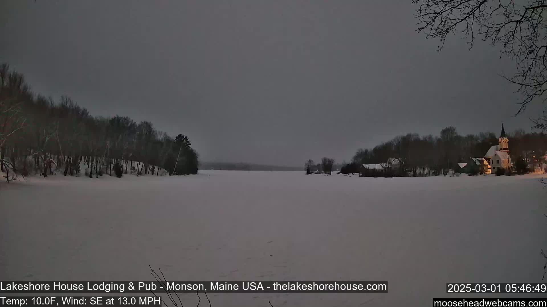 A snow-covered frozen lake is seen at dusk, with a building illuminated on the far shore and trees lining the banks under a dark gray sky, with a southeast wind at 13 mph and a temperature of 10°F.
