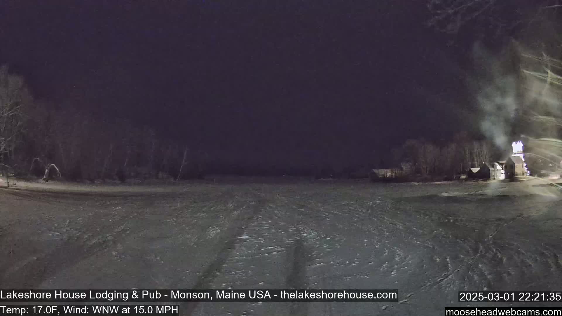 A snow-covered, dark landscape at night, under a dark sky, with a building and trees in the distance, and a 15 mph wind.