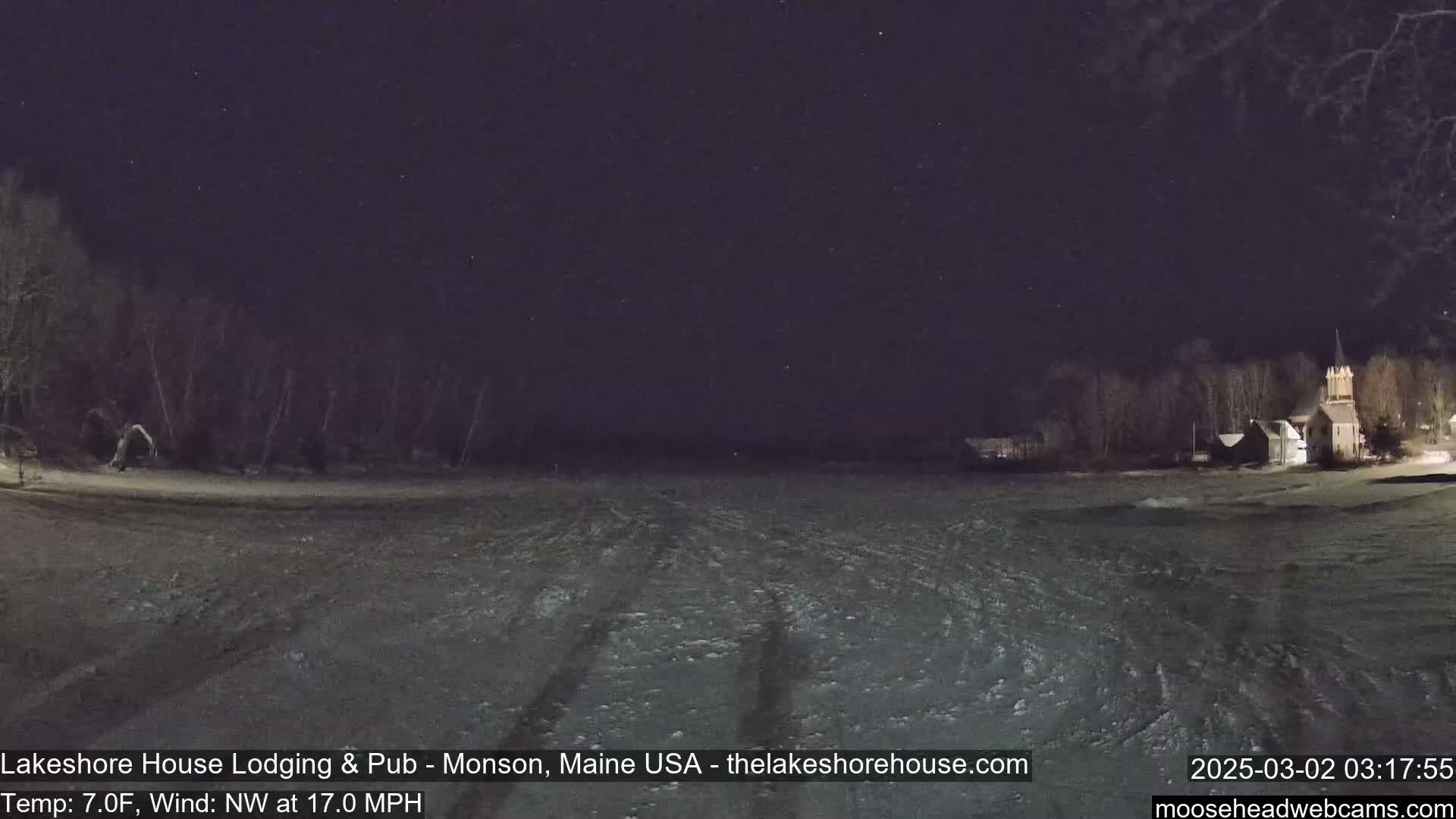 A snow-covered field at night under a starry sky, with trees and a building visible in the distance, a northwest wind blowing at 17 mph and a temperature of 7°F.