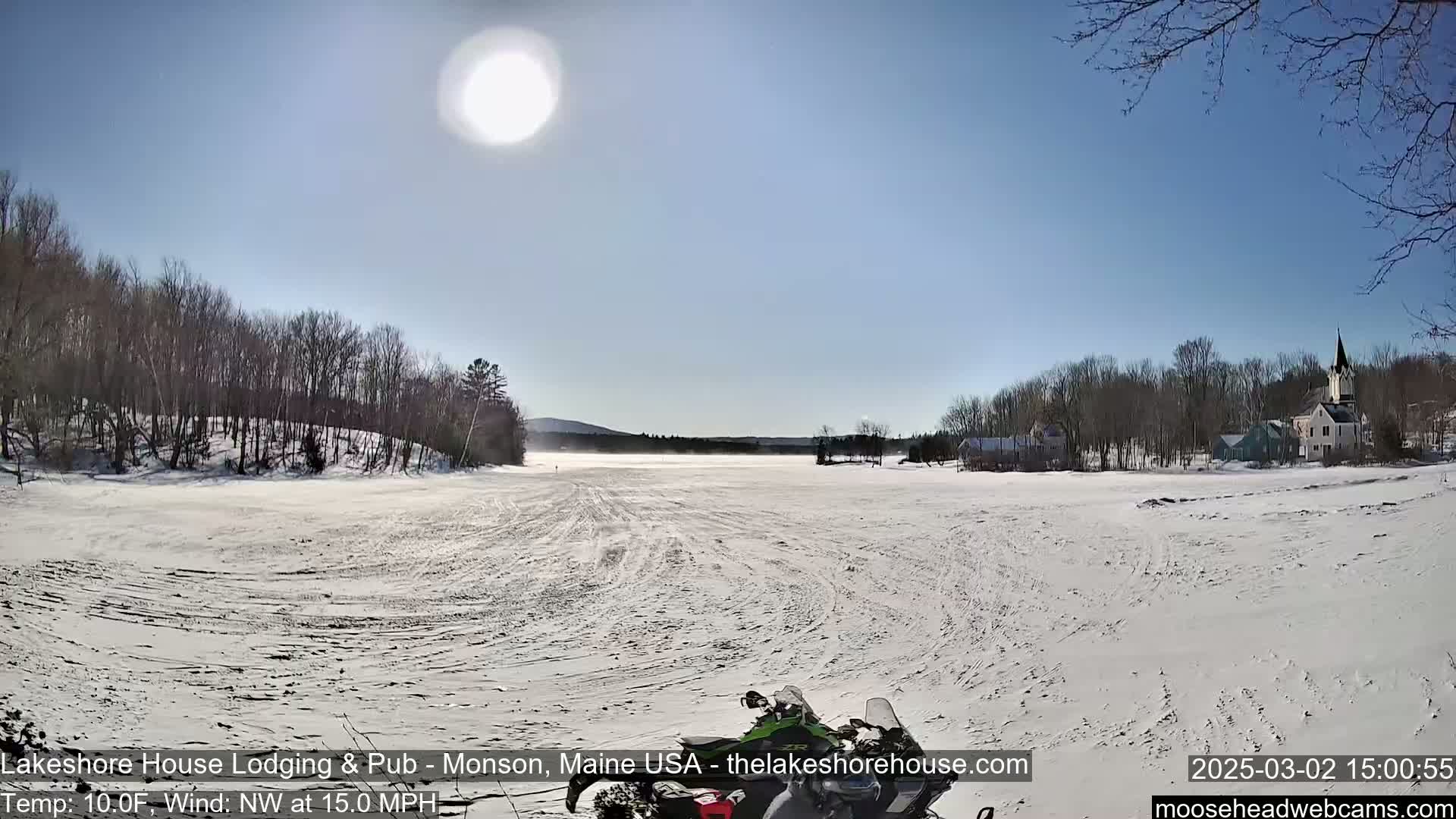 A snowmobile sits on a snowy, flat expanse of land, with a village and wooded hills in the background under a bright, sunny sky.