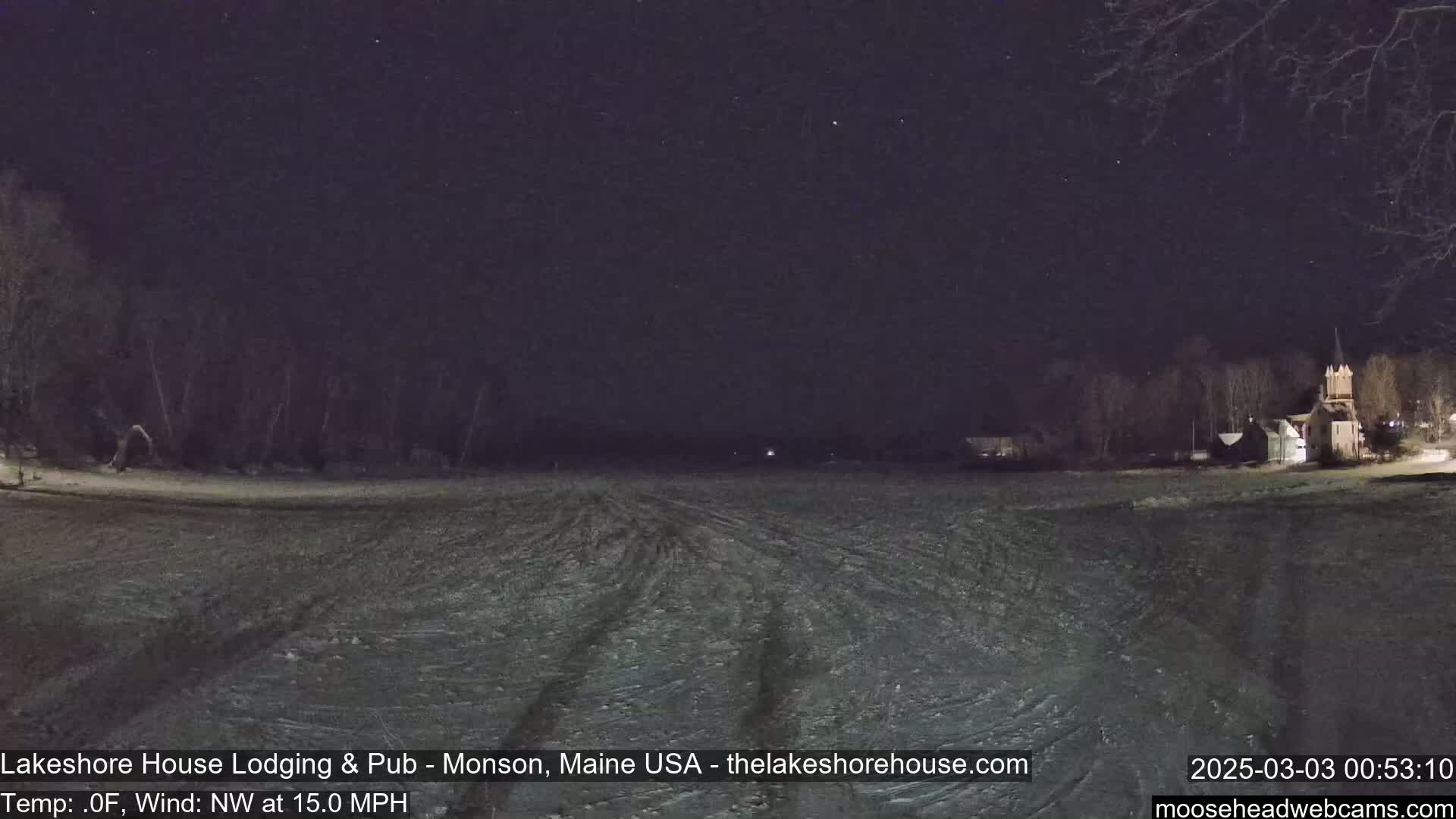 A snow-covered field is visible at night under a starry sky, with a building and trees in the distance, and a northwest wind blowing at 15 mph.
