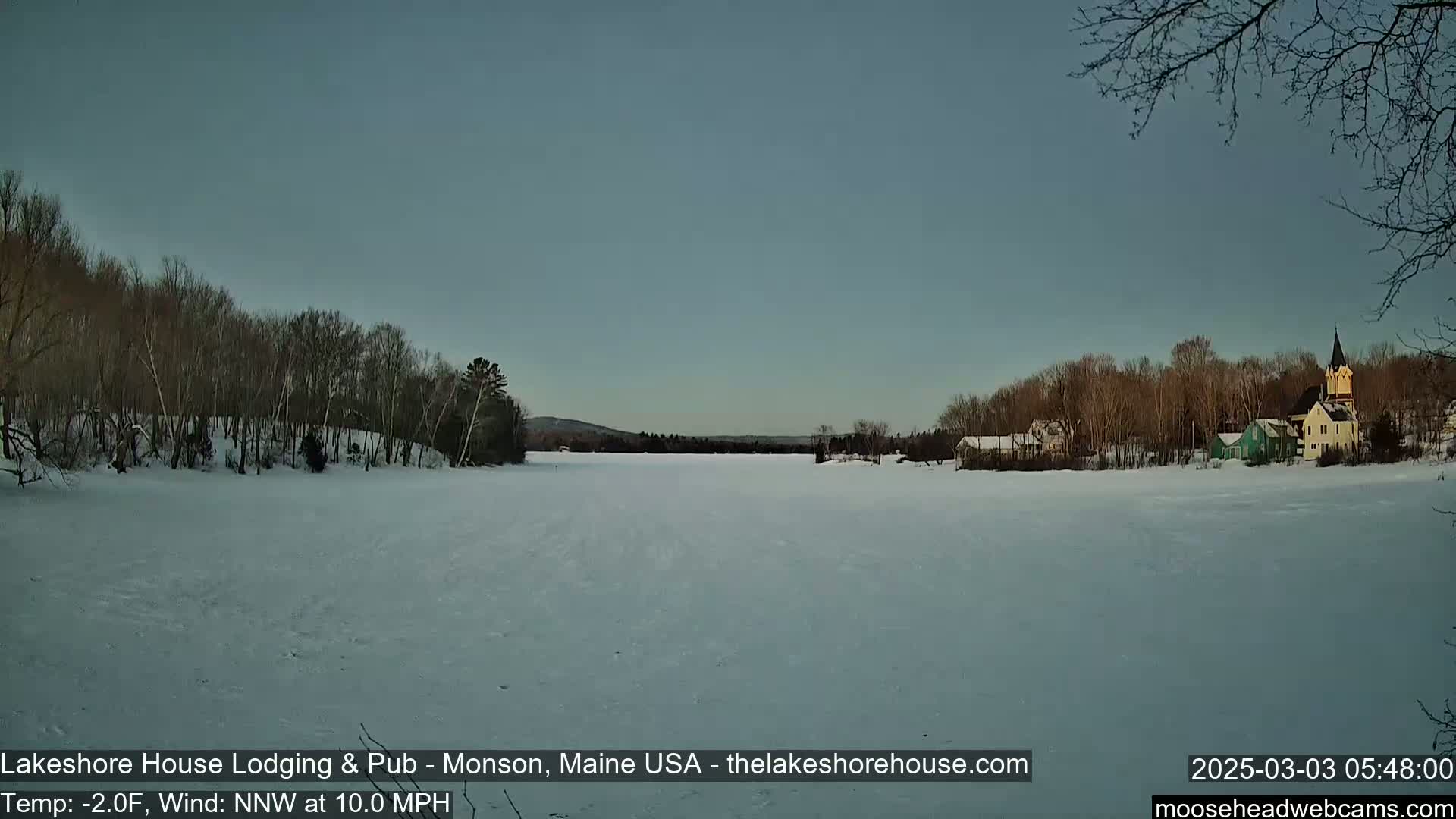A frozen lake stretches out before a line of snow-covered trees and buildings, under a pale, clear sky with a cold wind blowing.
