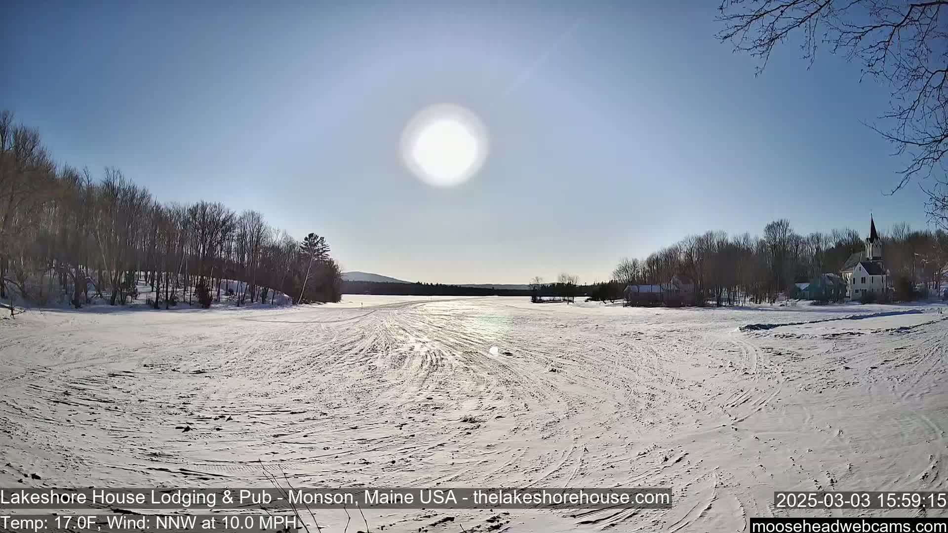 A snow-covered field stretches out to a frozen lake, with bare trees lining the shore and buildings in the distance under a bright, sunny sky.