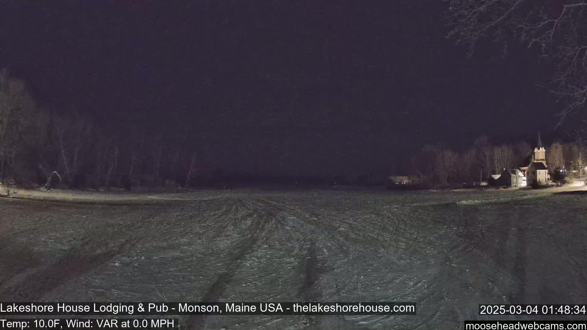 A nighttime, snow-covered field with a few buildings and trees visible in the distance under a dark sky.