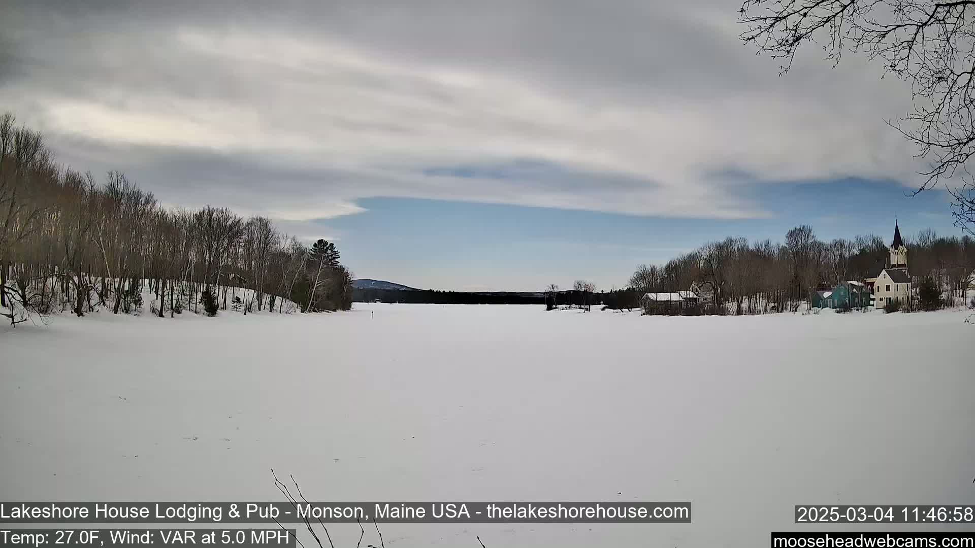 A snow-covered lake stretches toward a village under a partly cloudy sky.