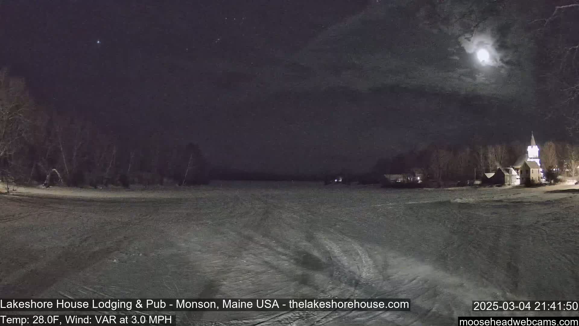 A nighttime view of a snow-covered landscape with a church steeple in the distance under a mostly cloudy sky with a bright moon.