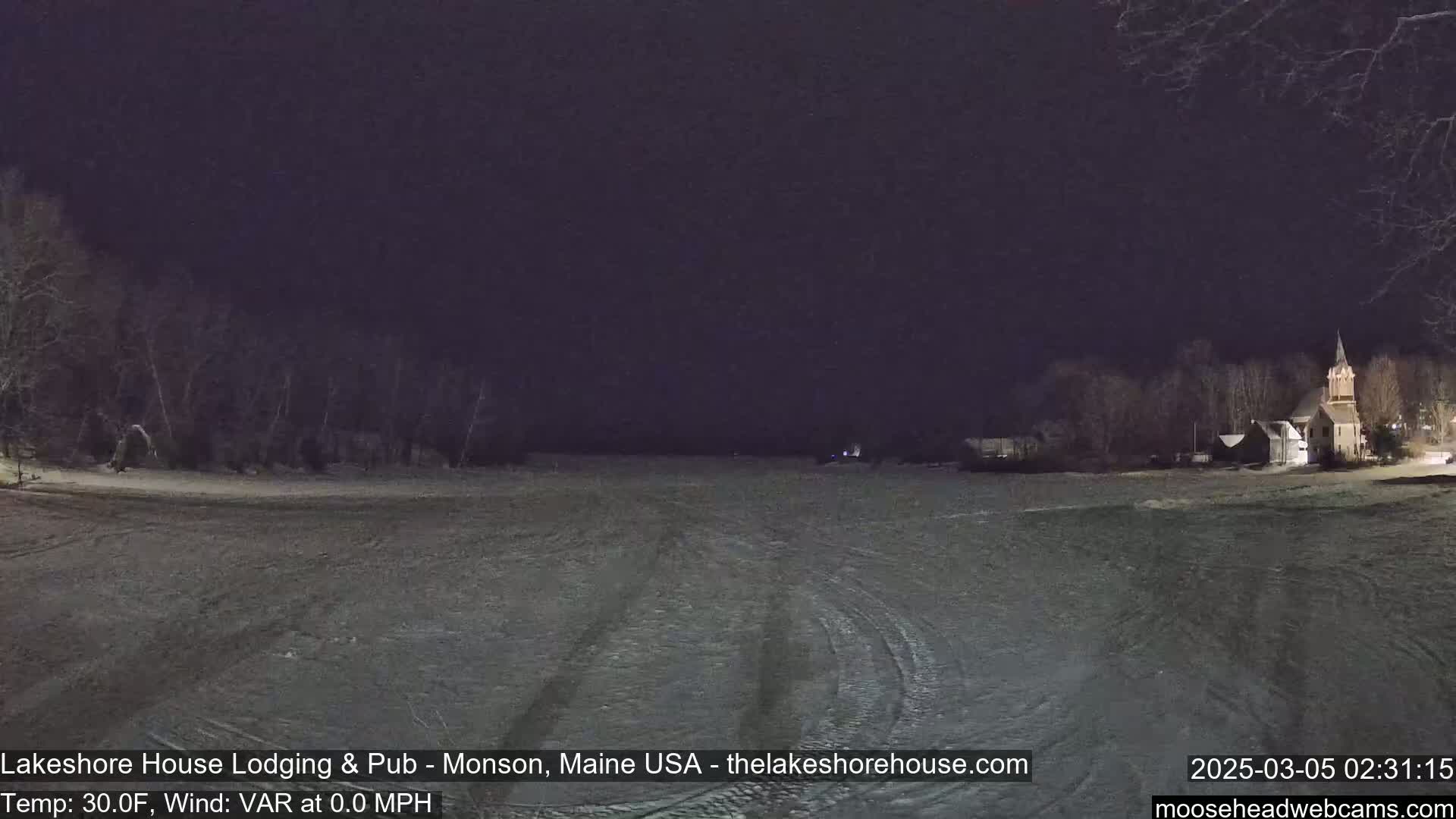 A nighttime view of a snow-covered landscape with a body of water and buildings in the distance under a dark sky.