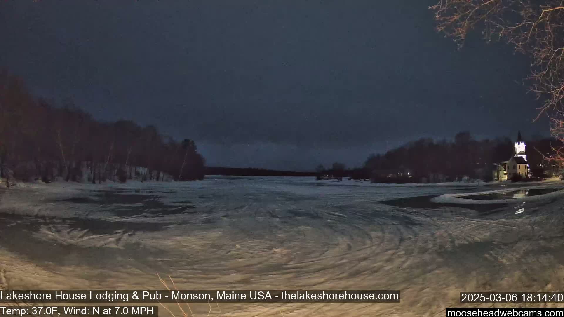 A partially frozen lake at night under a dark sky is seen from a lakeside vantage point, with a building illuminated in the distance and trees lining the shore.