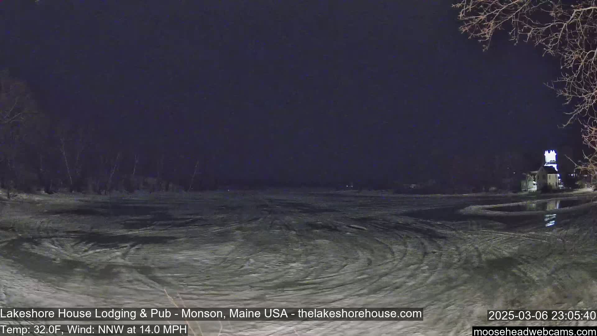 A nighttime view of a partially frozen body of water with swirling patterns in the ice and snow under a dark sky, with a building faintly visible in the distance.
