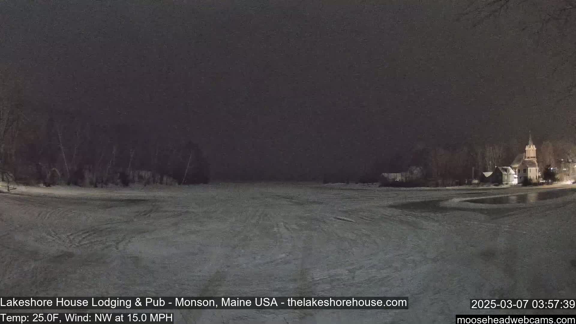 A snow-covered, frozen lake is seen at night, with a few buildings and trees visible in the distance under a dark sky.