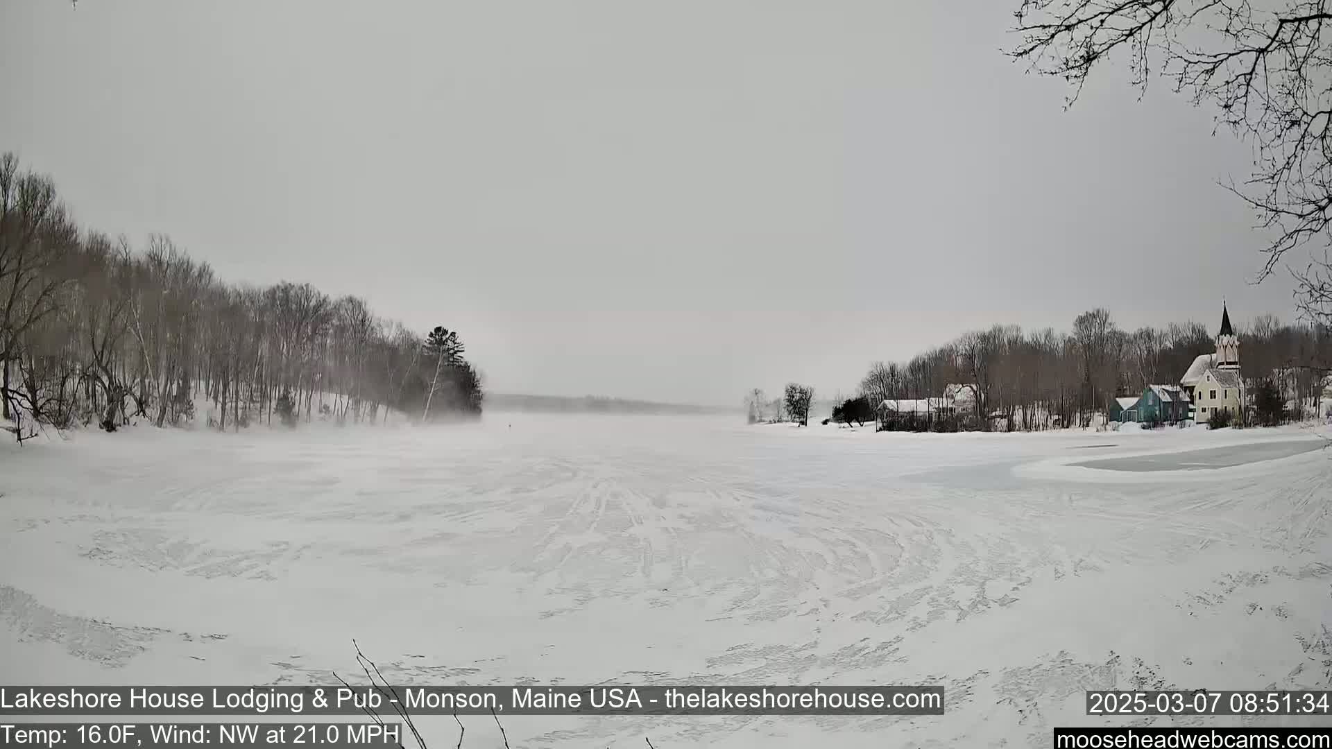 A snow-covered frozen lake stretches toward a small town with buildings and a church steeple under a gray, overcast sky, with a northwest wind blowing at 21 mph.