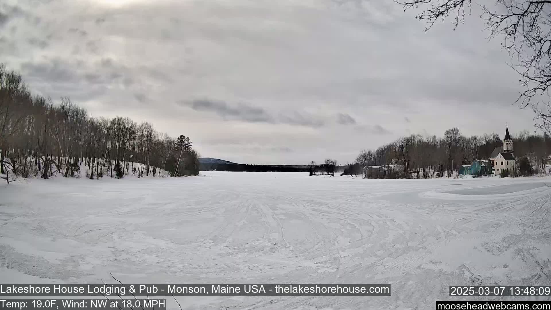 A frozen lake, under an overcast sky, shows tire tracks on its surface and a small town with bare trees in the background.