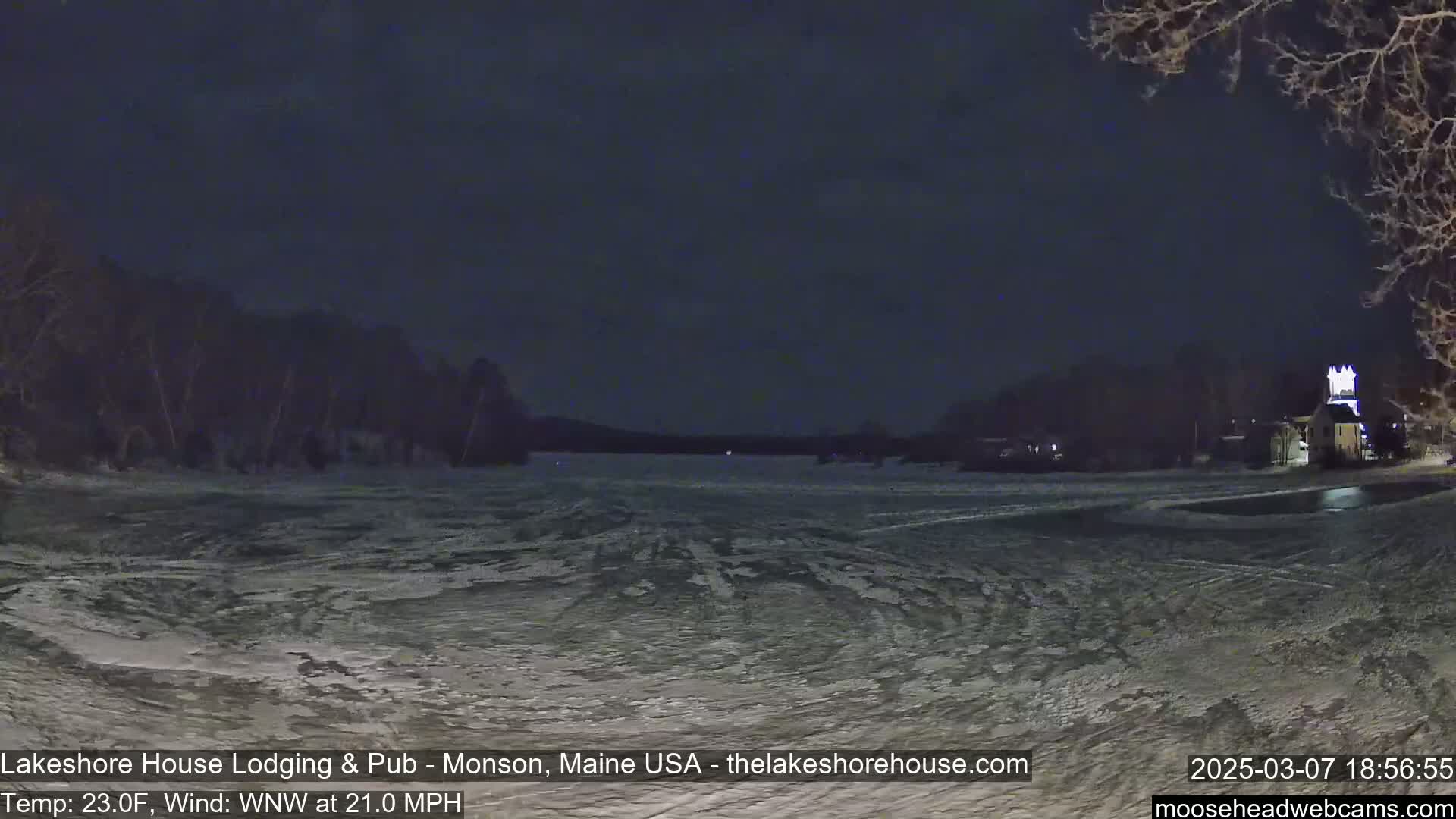 A frozen lake at night under a dark sky is seen from across a snowy shore, with buildings and trees visible in the distance under a WNW wind of 21 mph and 23°F temperature.