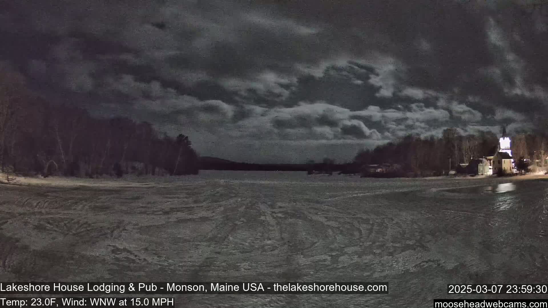 A partially frozen lake is seen at night under a cloudy sky, with snow-covered trees and a lit building visible on the shoreline.