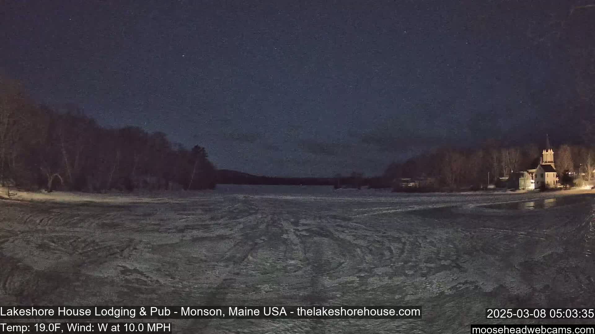 A frozen lake at night under a dark sky is viewed from across the water, showing a shoreline with buildings and trees.