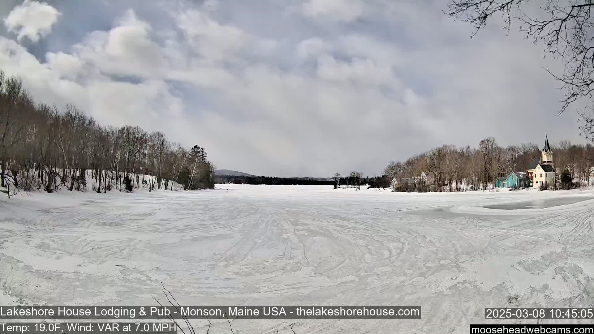 A frozen lake, covered in snow and crisscrossed with tracks, stretches out before a small village nestled among bare, winter trees under a partly cloudy sky.