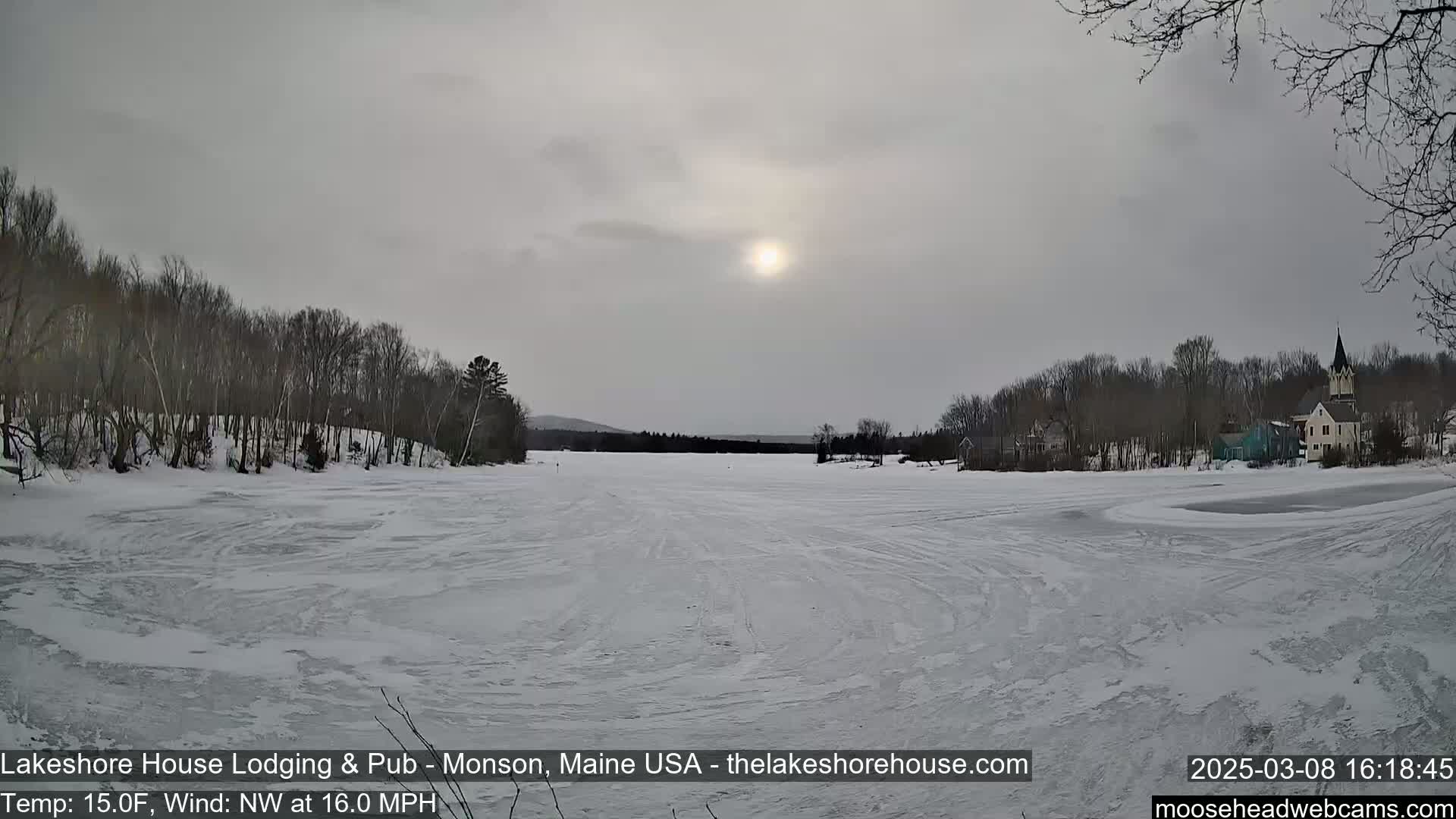 A frozen lake, under a mostly cloudy sky with a visible sun, is surrounded by bare trees and a small town in the distance.