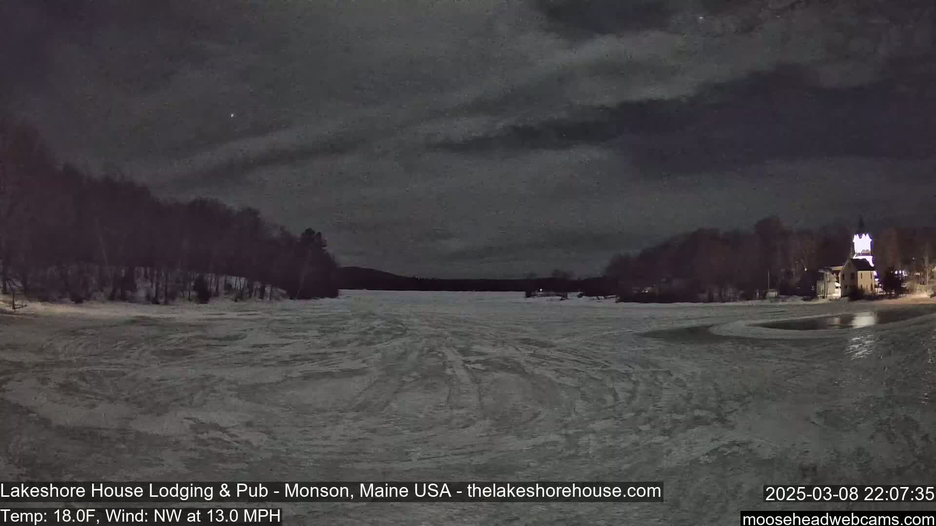 A frozen lake at night under a mostly cloudy sky is viewed from across a snowy shoreline, with a building lit up on the far right.