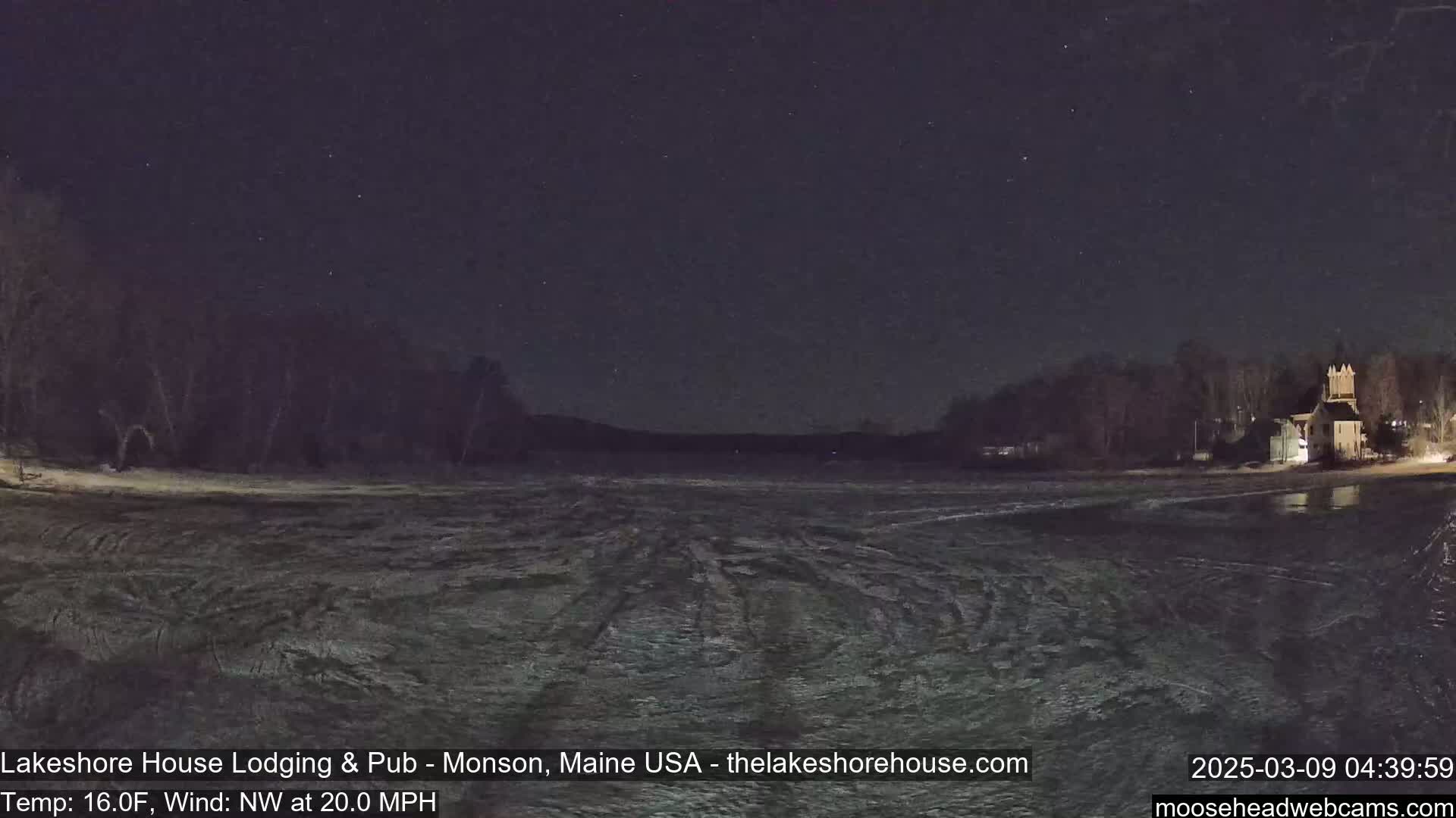 A nighttime view of a frozen lake under a starry sky, with a building and trees visible on the far shore, and a 16°F temperature with a 20 mph northwest wind.