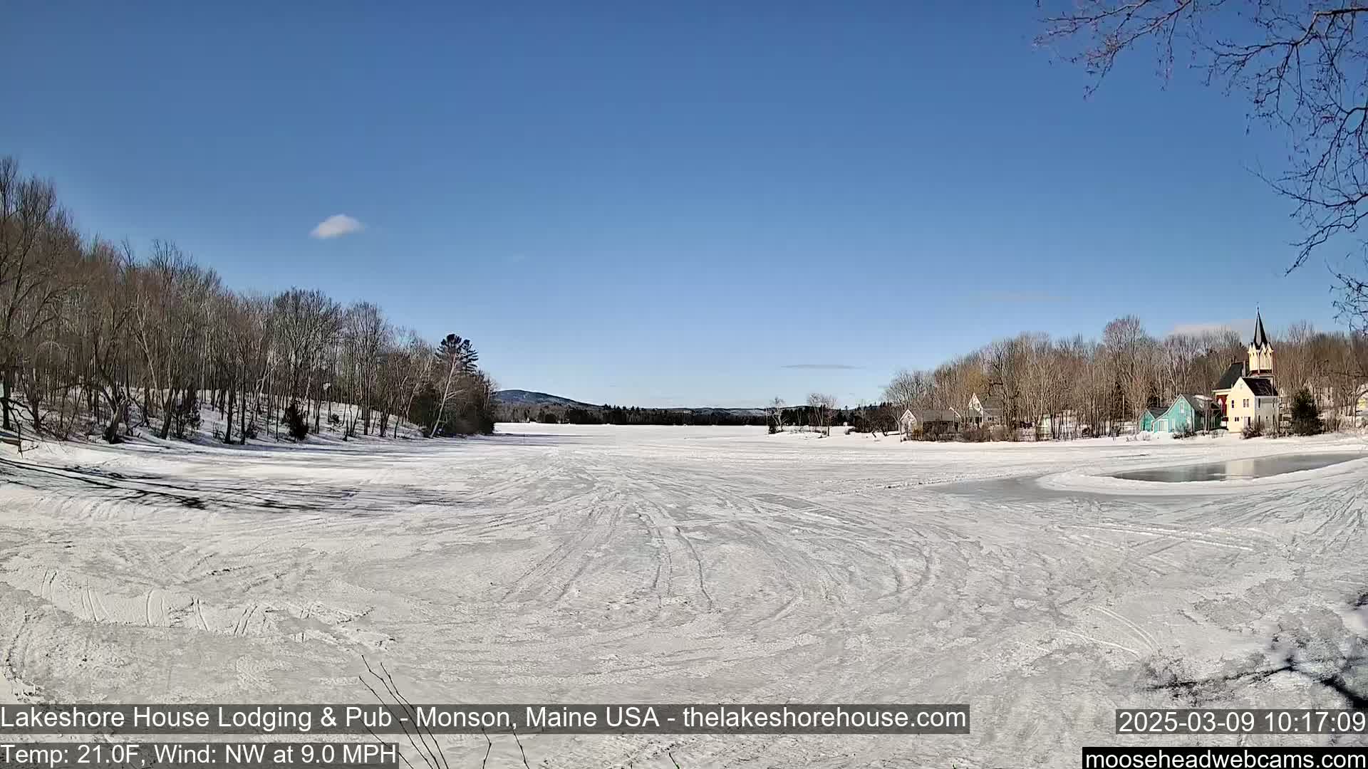 A frozen lake with tire tracks across its surface is surrounded by snow-covered trees and a small village under a clear blue sky.
