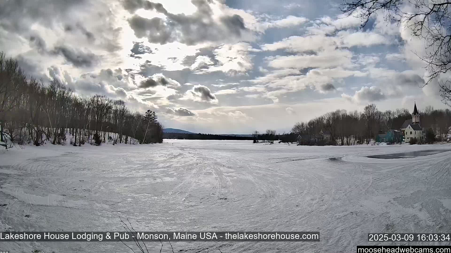 A partly cloudy winter day shows a frozen lake with tire tracks, bare trees lining the shore, and buildings in the distance.