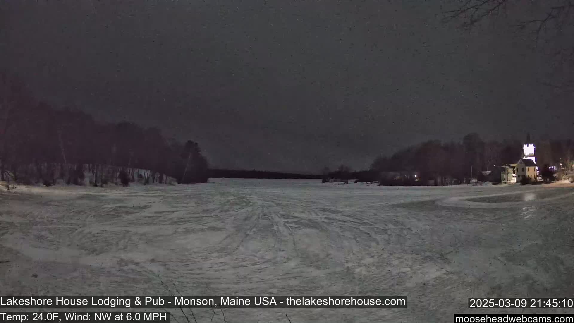 A snow-covered frozen lake is seen at night under a dark sky, with a building lit up in the distance and trees lining the shore.
