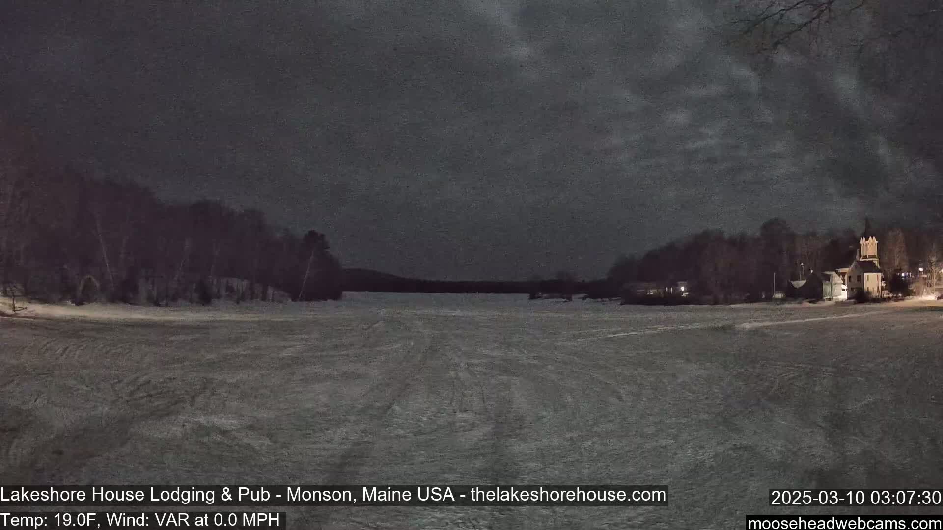 A snow-covered landscape at night, under a dark sky, shows a frozen lake with buildings and trees in the distance.