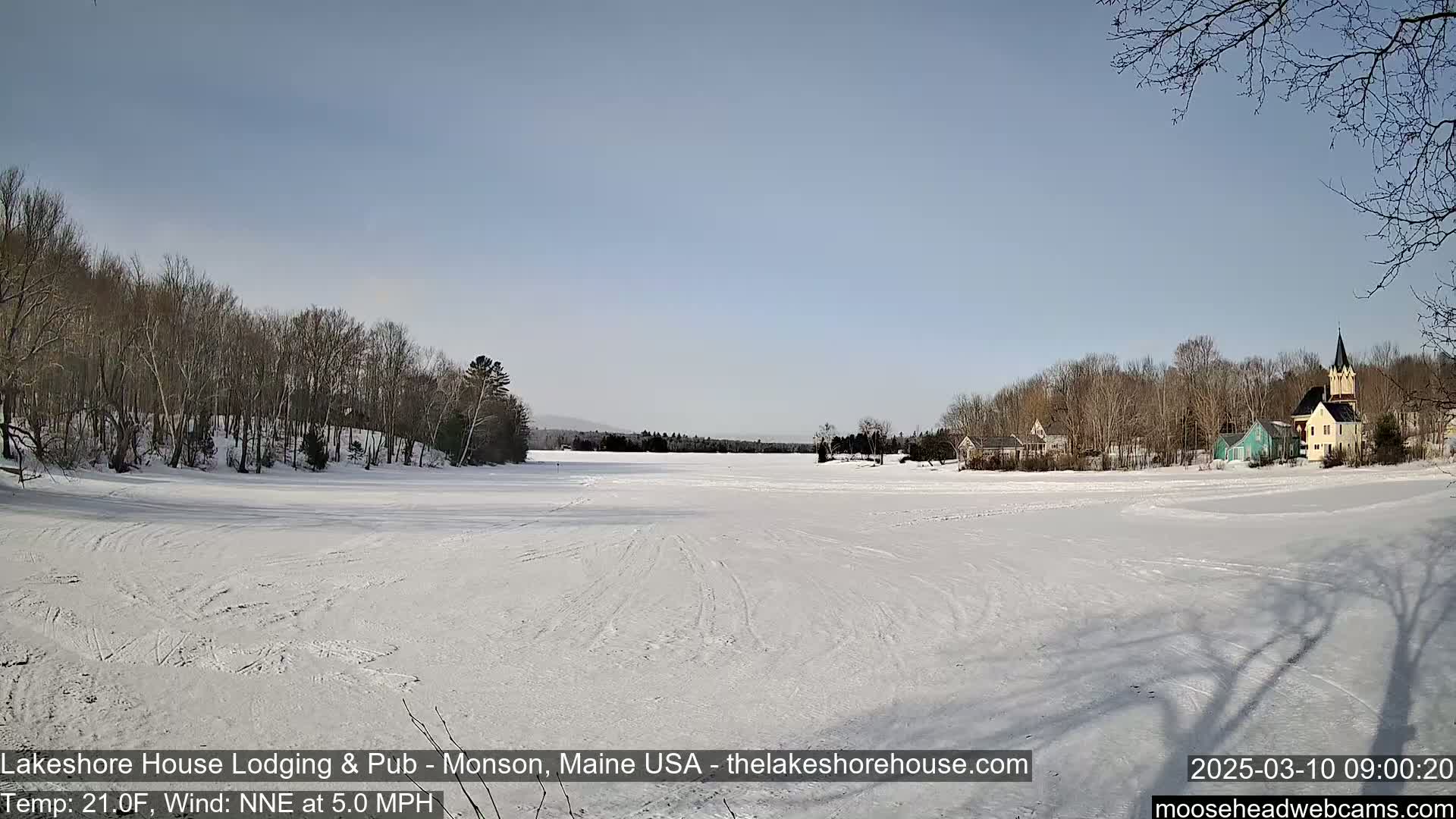 A snow-covered frozen lake stretches out before a small village of houses and a church under a clear blue sky.