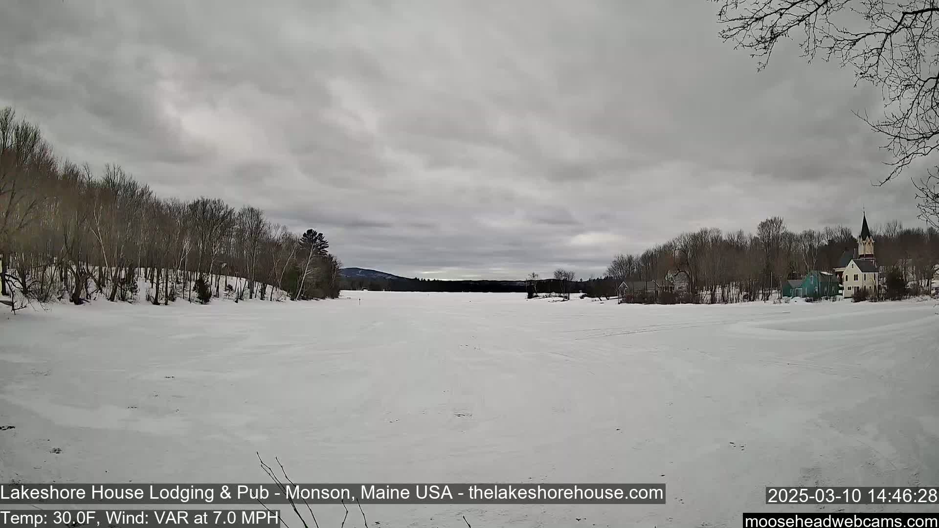 A snow-covered frozen lake stretches to a shoreline of bare trees and buildings under a gray, overcast sky.