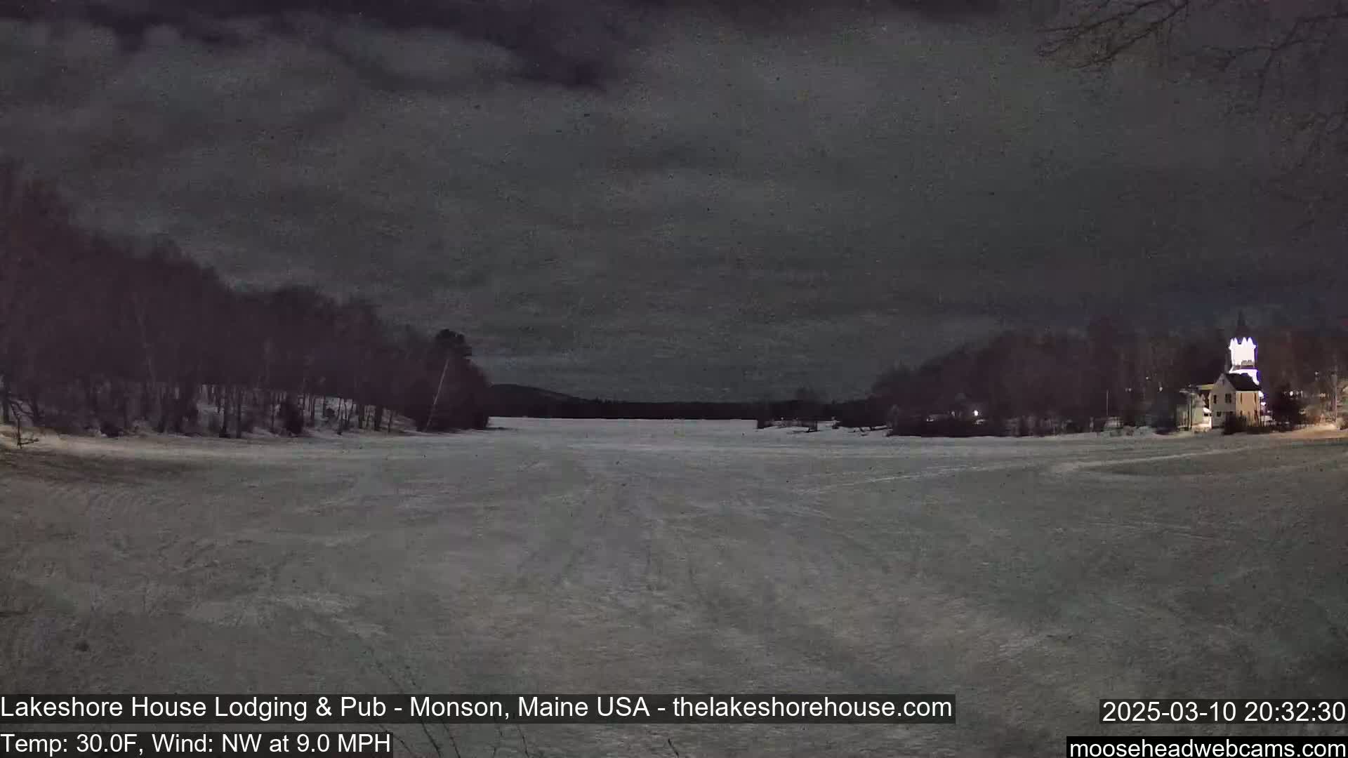 A snow-covered frozen lake is seen at night under a dark, cloudy sky, with trees and a building lit up in the distance.