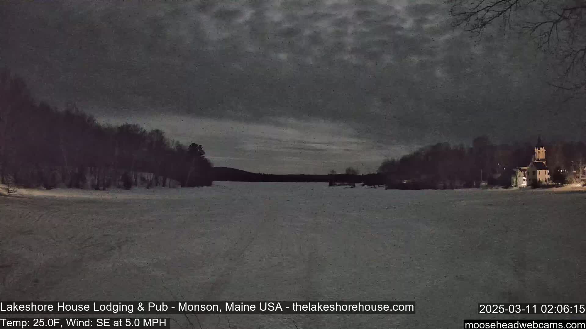 A snow-covered frozen lake is seen at night under a cloudy sky, with dark trees lining the shore and a building faintly lit in the distance.