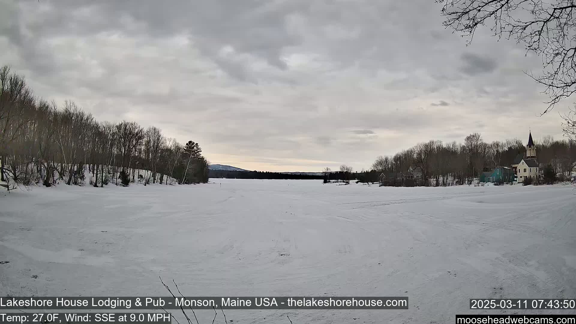 A snow-covered frozen lake stretches out to a village of buildings and trees under a cloudy sky.
