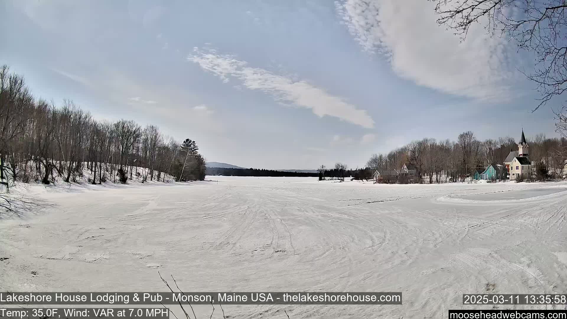 A snow-covered frozen lake stretches before a line of trees and buildings under a partly cloudy sky.