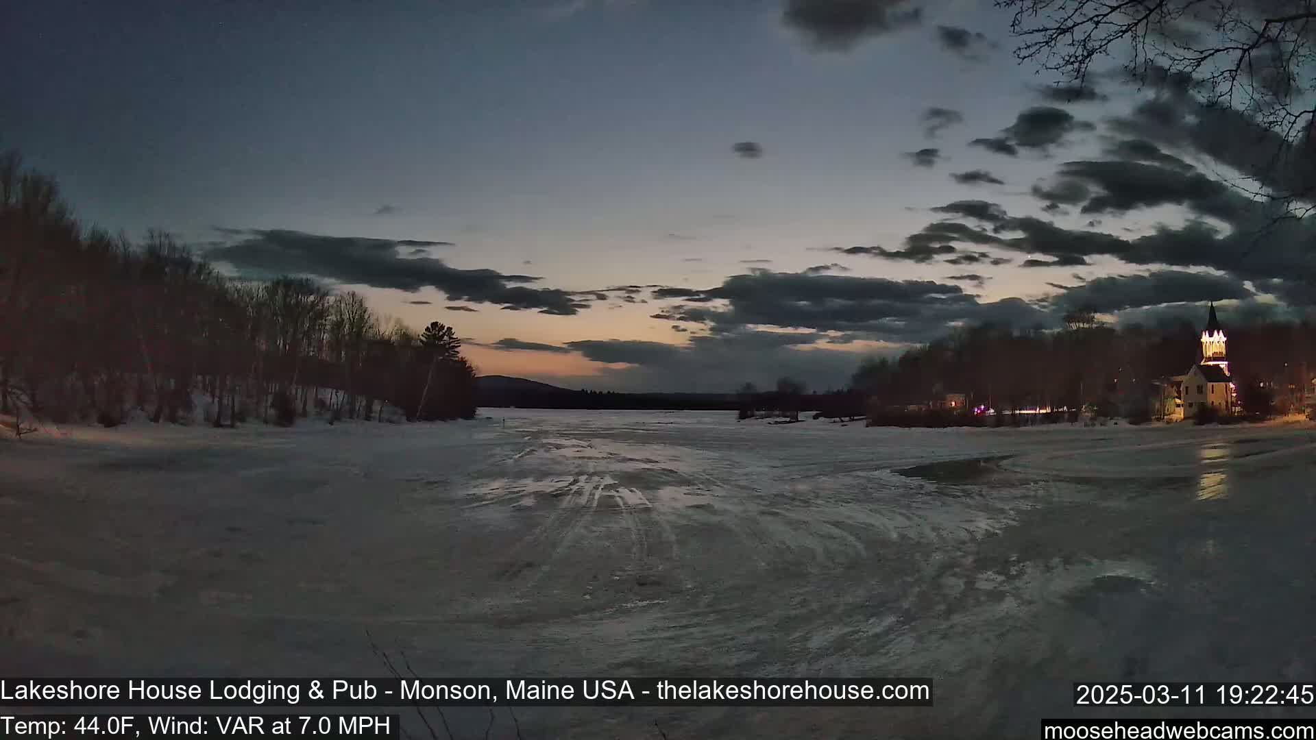 A partially frozen lake at dusk, under a partly cloudy sky, with a lit-up building visible on the far shore and trees lining the edges.