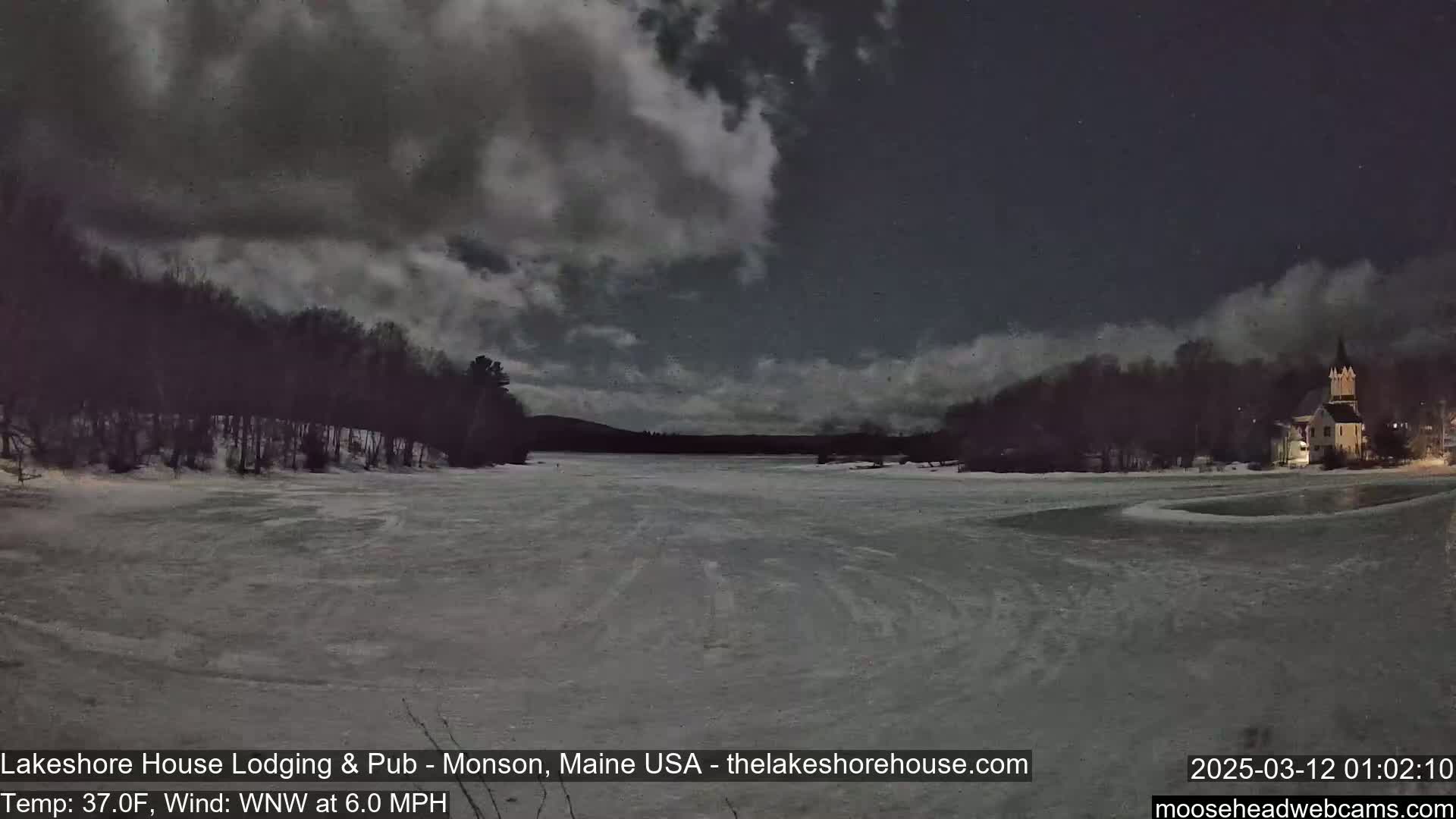 A frozen lake under a partly cloudy night sky is seen, with a building and trees visible in the distance.